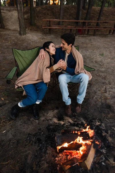Smiling interracial couple holding hands on chairs near campfire — Stock Photo