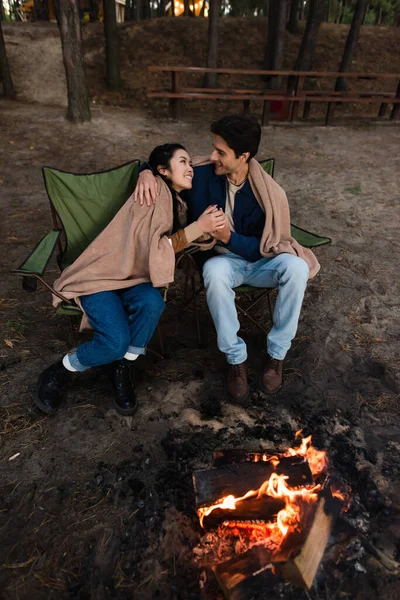 Sonriente pareja interracial cogida de la mano en sillas cerca de fogata - foto de stock