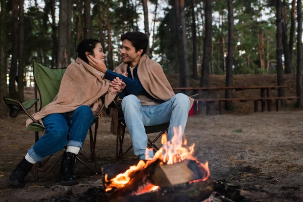 Smiling man touching face of asian girlfriend near blurred campfire outdoors — Stock Photo