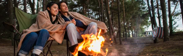 Smiling multiethnic couple in blankets sitting near blurred campfire, banner — Stock Photo