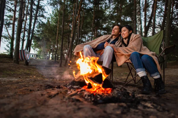 Jeune couple multiethnique dans des couvertures assis près du feu de camp flou — Photo de stock