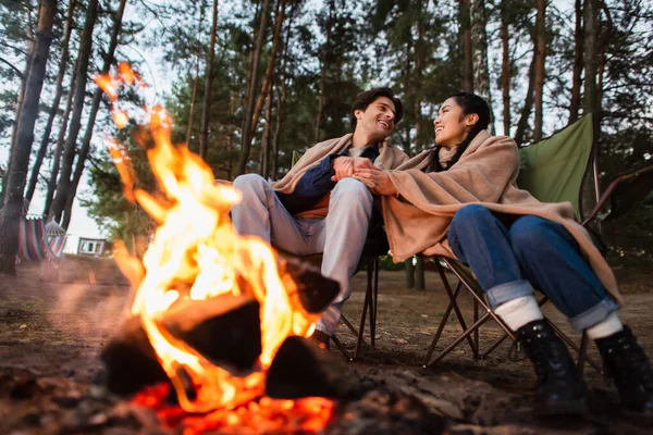 Multiethnic couple holding hands near blurred campfire during weekend — Stock Photo