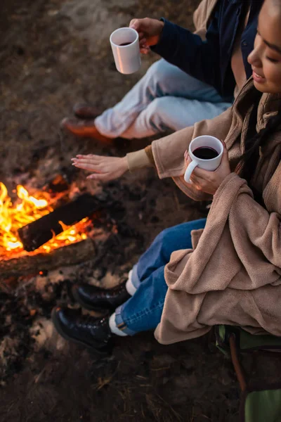 High angle view of smiling asian woman holding cup near boyfriend and campfire — Stock Photo