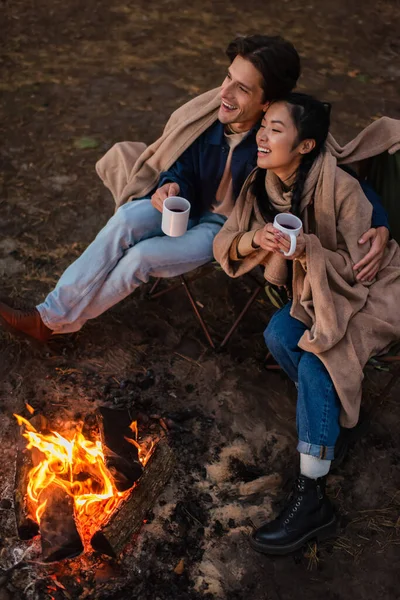 Улыбающаяся межрасовая пара с чашками, обнимающимися у костра в выходные — стоковое фото