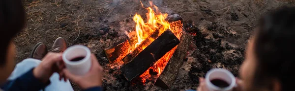 Lagerfeuer neben verschwommenem Paar mit Tassen im Freien, Banner — Stockfoto