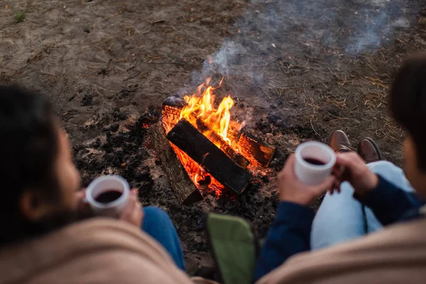 Vue grand angle du feu de camp près d'un couple flou avec des tasses de café à l'extérieur — Photo de stock