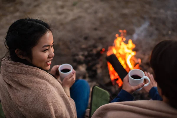 Vue grand angle de sourire femme asiatique avec tasse floue regardant petit ami près de feu de camp — Photo de stock
