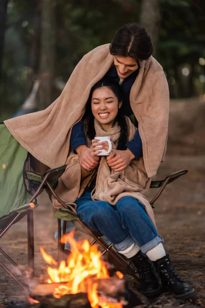 Mann hält Hand von positiver asiatischer Freundin mit Tasse am verschwommenen Lagerfeuer — Stockfoto