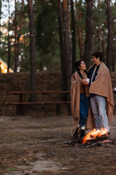 Couple interracial dans des couvertures avec tasse parler près de feu de camp dans la soirée — Photo de stock