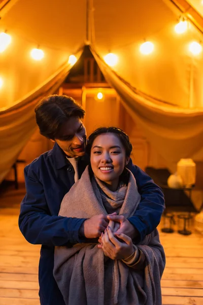 Mann umarmt glückliche asiatische Freundin in Decke nahe Glamping-Haus mit Beleuchtung im Freien — Stockfoto