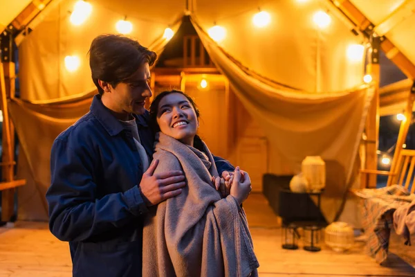 Man embracing asian girlfriend in blanket near blurred glamping house during weekend — Stock Photo