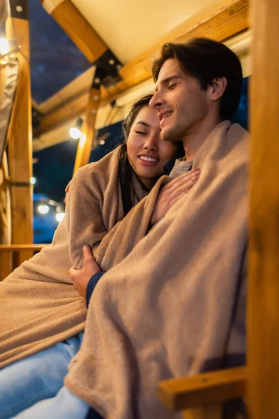 Sonriendo mujer asiática en manta abrazando novio en glamping terraza - foto de stock