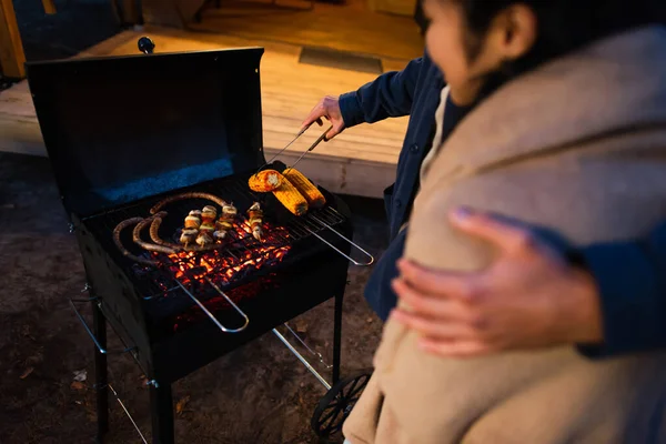 Cropped view of man cooking on grill and hugging blurred girlfriend outdoors — Stock Photo