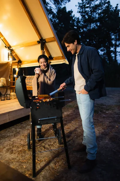 Smiling interracial couple with cup cooking on grill on glamping terrace — Stock Photo