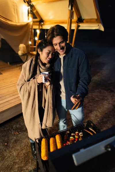 Multiethnic couple with cup grilling food near glamping house — Stock Photo