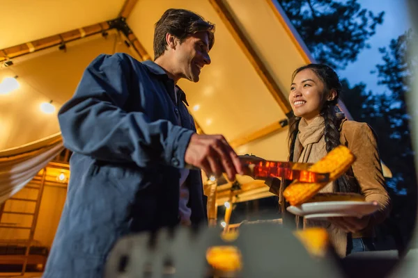 Low angle view of smiling interracial couple cooking on grill near glamping house in evening — Stock Photo