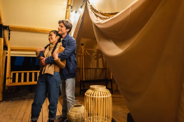 Multiethnic couple hugging on terrace of glamping house with light garland — Stock Photo