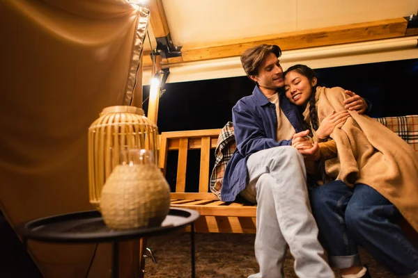 Мультиэтническая пара с одеялом, держащаяся за руки на скамейке возле глампинг-хауса — стоковое фото