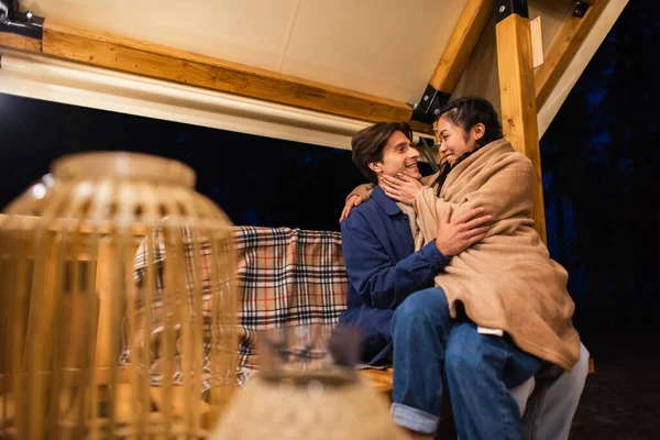 Side view of asian woman in blanket touching boyfriend on bench near glamping house — Stock Photo