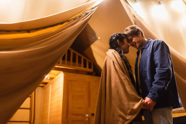 Улыбающиеся мультиэтнические пары держатся за руки возле светлых гирлянд и глампинг-хауса — стоковое фото