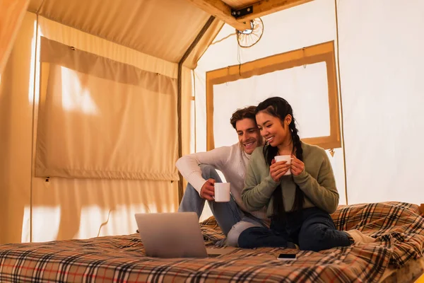 Sorridente coppia interrazziale con tazze guardare film sul computer portatile sul letto in glamping casa — Foto stock