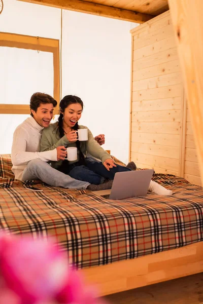 Excited asian woman pointing at laptop near boyfriend with cup on bed in glamping house — Stock Photo