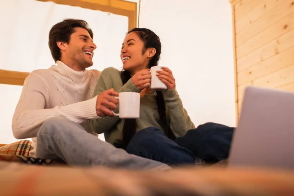 Laughing interracial couple with cups sitting near blurred laptop in glamping house — Stock Photo