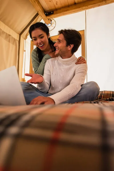 Souriant homme pointant vers ordinateur portable près de petite amie asiatique dans la maison glamping — Photo de stock