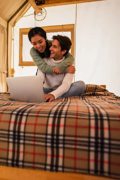 Cheerful asian woman hugging boyfriend freelancer on bed in glamping house — Stock Photo