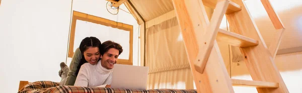 Fröhliches gemischtrassiges Paar mit Laptop im Glamping-Haus auf dem Bett liegend, Banner — Stockfoto