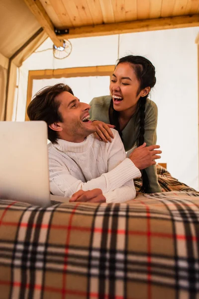 Excited interracial couple laughing near blurred laptop on bed in glamping house — Stock Photo