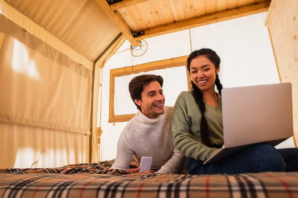 Multiethnic couple using gadgets on bed in glamping house — Stock Photo