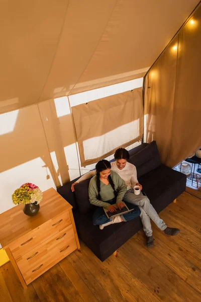 Vista de ángulo alto de la pareja interracial con taza usando el ordenador portátil en la casa de glamping - foto de stock