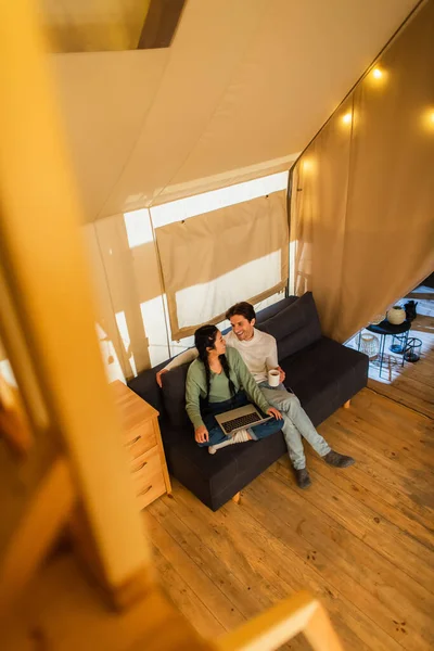 Visão de alto ângulo de casal interracial com copo e laptop sentado no sofá na casa glamping — Fotografia de Stock
