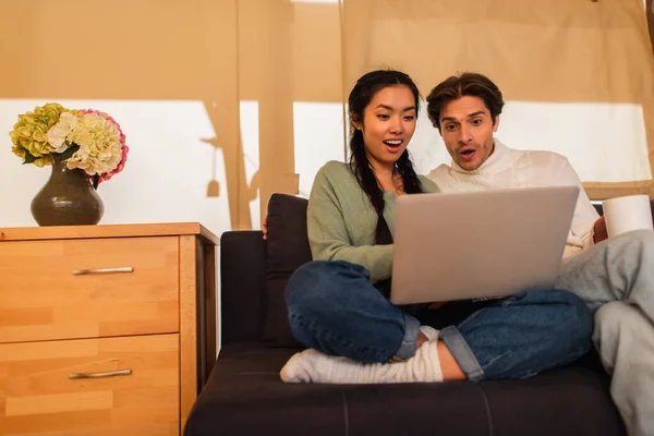 Stupito coppia interrazziale con tazza utilizzando computer portatile sul divano in glamping casa — Foto stock