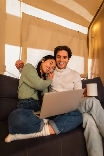 Sorrindo casal multiétnico com copo e laptop sentado no sofá em casa glamping — Fotografia de Stock