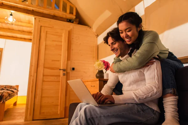 Sorridente donna asiatica abbracciare fidanzato con computer portatile sul divano in glamping casa — Foto stock