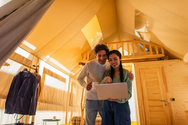 Man with cup hugging asian girlfriend pointing at laptop in glamping house — Stock Photo