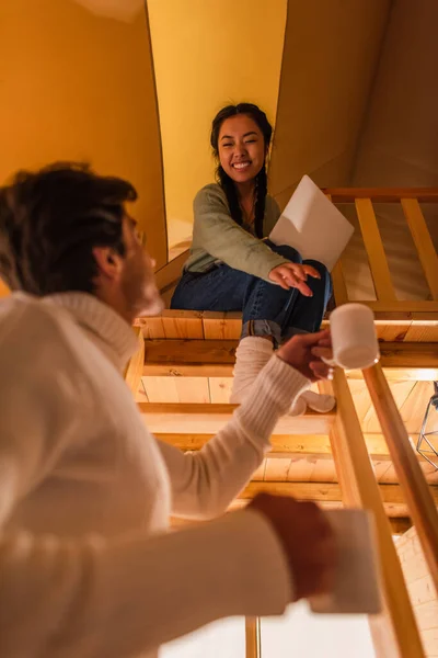 Vue à angle bas de l'homme tenant des tasses près de petite amie asiatique avec ordinateur portable dans la maison de glamping — Photo de stock