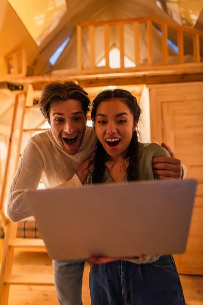 Amazed man hugging asian girlfriend with laptop in glamping house — Stock Photo