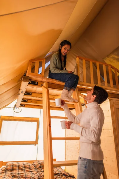 Smiling man holding cups near asian girlfriend with laptop in glamping house — Stock Photo