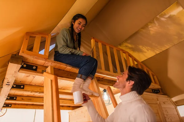 Low angle view of smiling asian woman with laptop looking at boyfriend with cup in glamping house — Stock Photo