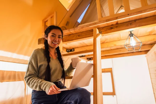 Asian woman with cup and laptop looking at camera in glamping house — Stock Photo