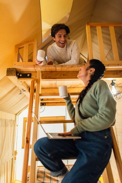Vista de ángulo bajo de la sonriente pareja interracial con tazas y portátil mirándose en la casa glamping - foto de stock
