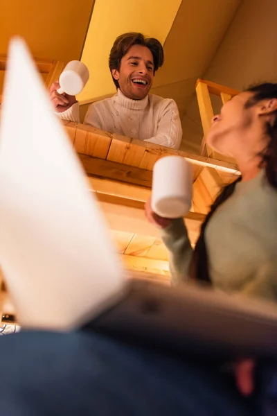 Low angle view of smiling man with cup looking at blurred girlfriend with laptop in glamping house — Stock Photo