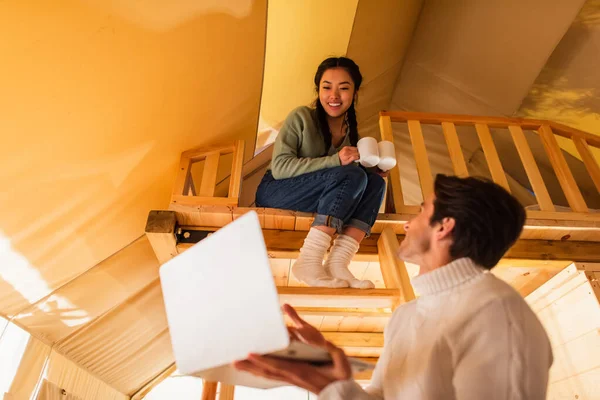 Cheerful asian woman holding cups near blurred boyfriend with laptop in glamping house — Stock Photo