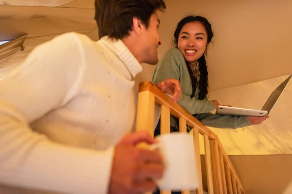 Smiling interracial couple with cup and laptop spending time in glamping house — Stock Photo