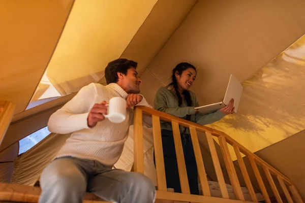 Low angle view of asian freelancer using laptop near boyfriend with cup in glamping house — Stock Photo