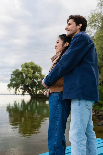 Vue latérale du jeune couple multiethnique embrassant près du lac — Photo de stock