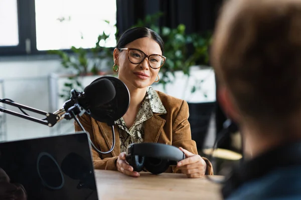 Sonriente asiático locutor en gafas celebración auriculares y mirando borrosa colega - foto de stock